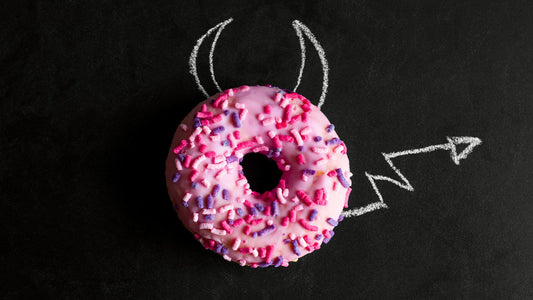 A bright, pink-iced donut with sprinkles sits against a black backdrop with chalked devil horns and tail.