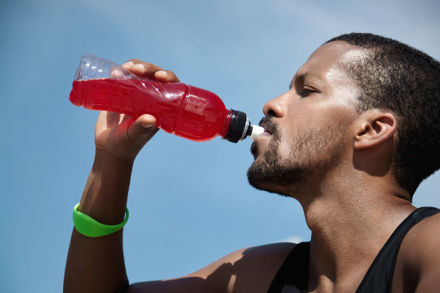 An athletic man drinks a red electrolyte beverage from a water bottle after working-out in the streams of sunlight. 